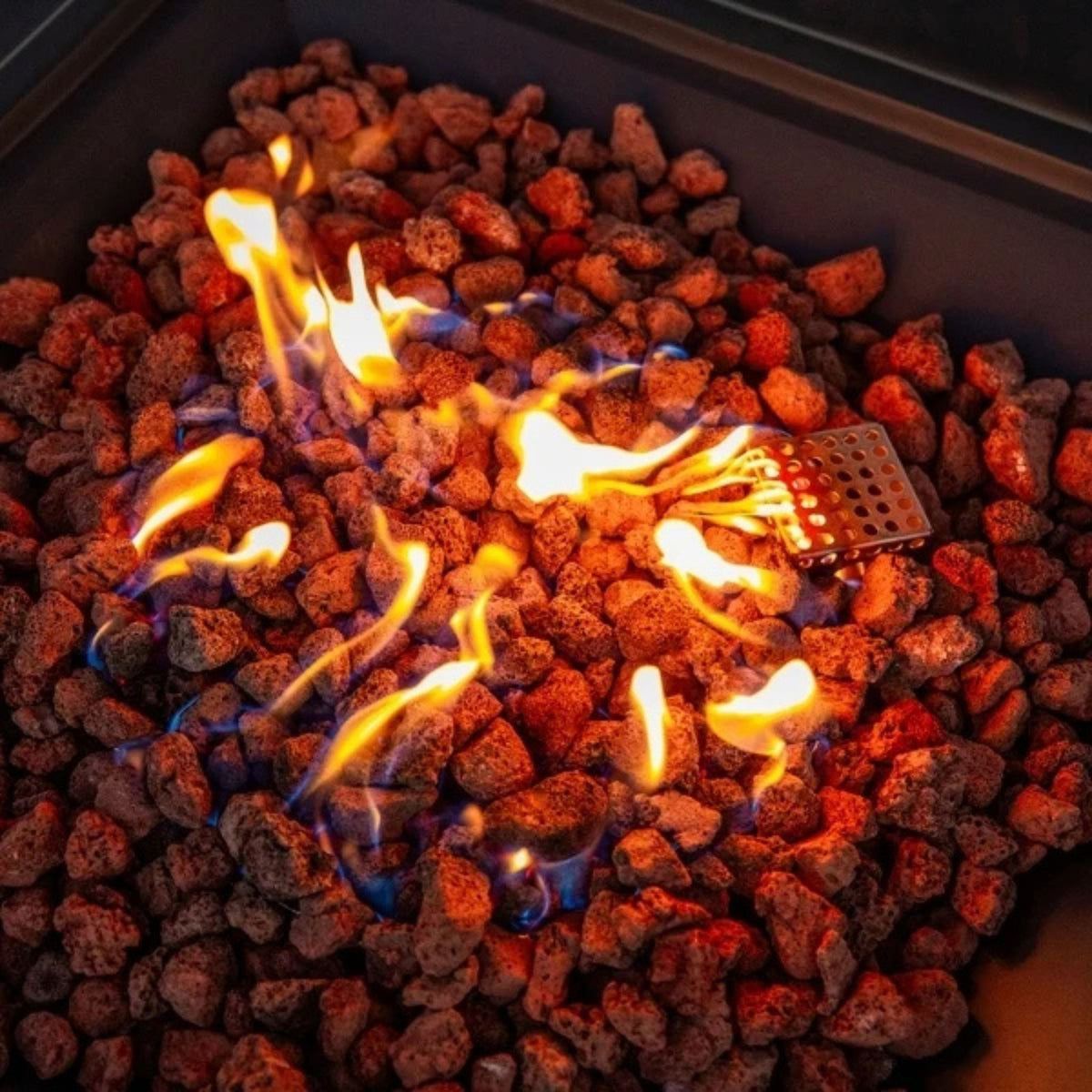 Close-up of a fire pit filled with glowing lava rocks and bright orange flames, with a small metal grate visible among the stones.