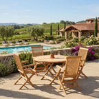 A round wooden outdoor dining table with four matching slatted folding chairs on a stone patio, set beside a swimming pool with a villa, vineyard, and hills in the background.