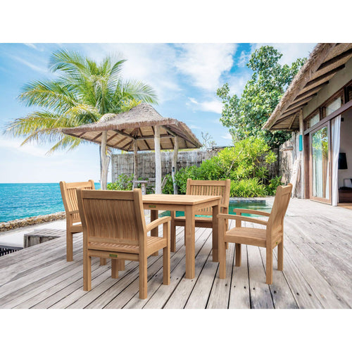 A wooden outdoor dining set with a rectangular table and four matching armchairs placed on a light deck beside a pool, with palm trees, ocean water, and a thatched umbrella in the background.