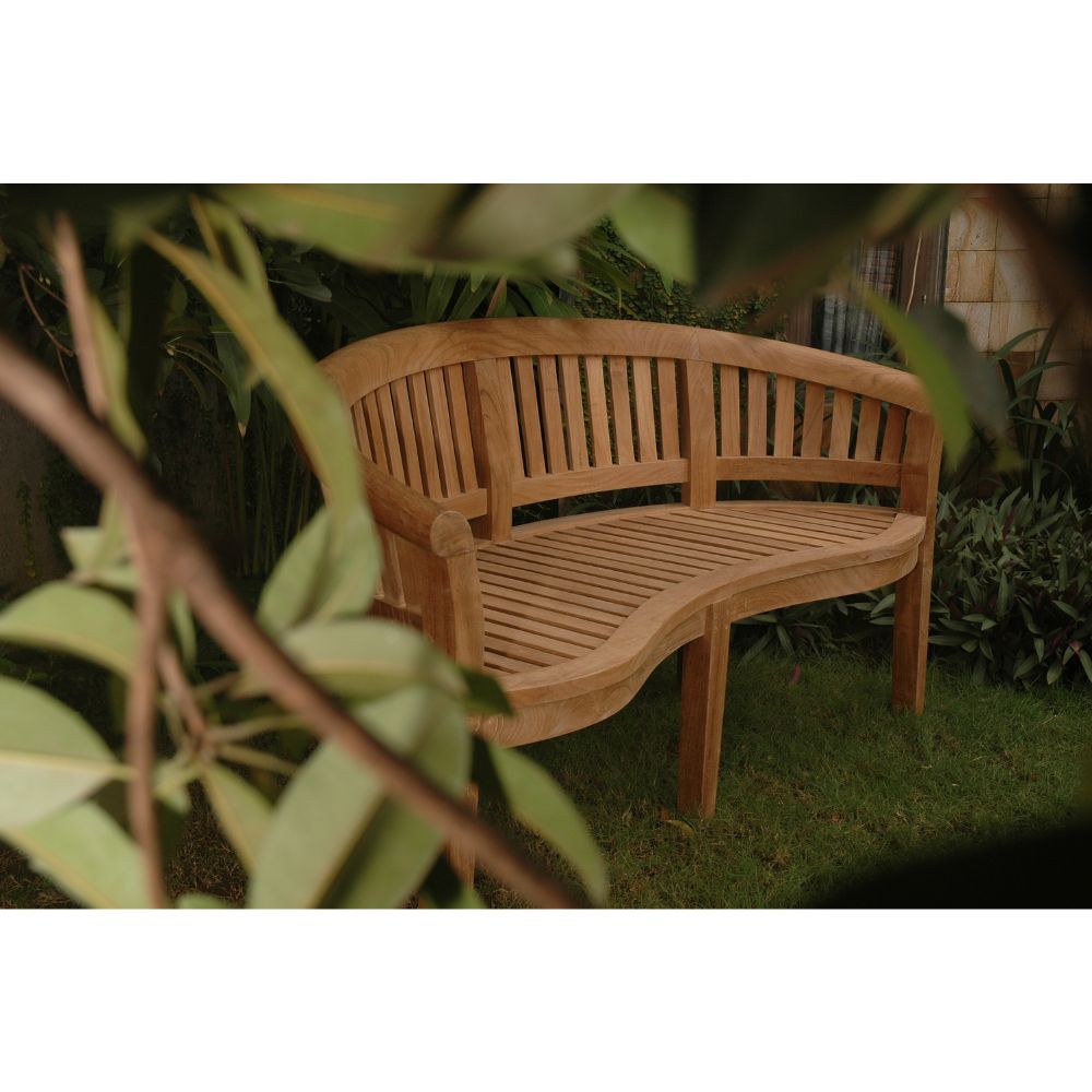 A curved wooden garden bench made of natural teak wood, set on a grassy lawn with green foliage in the background and foreground.