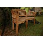 A curved wooden garden bench with a slatted back and seat, placed on grass in an outdoor garden setting with plants and a stone wall in the background.