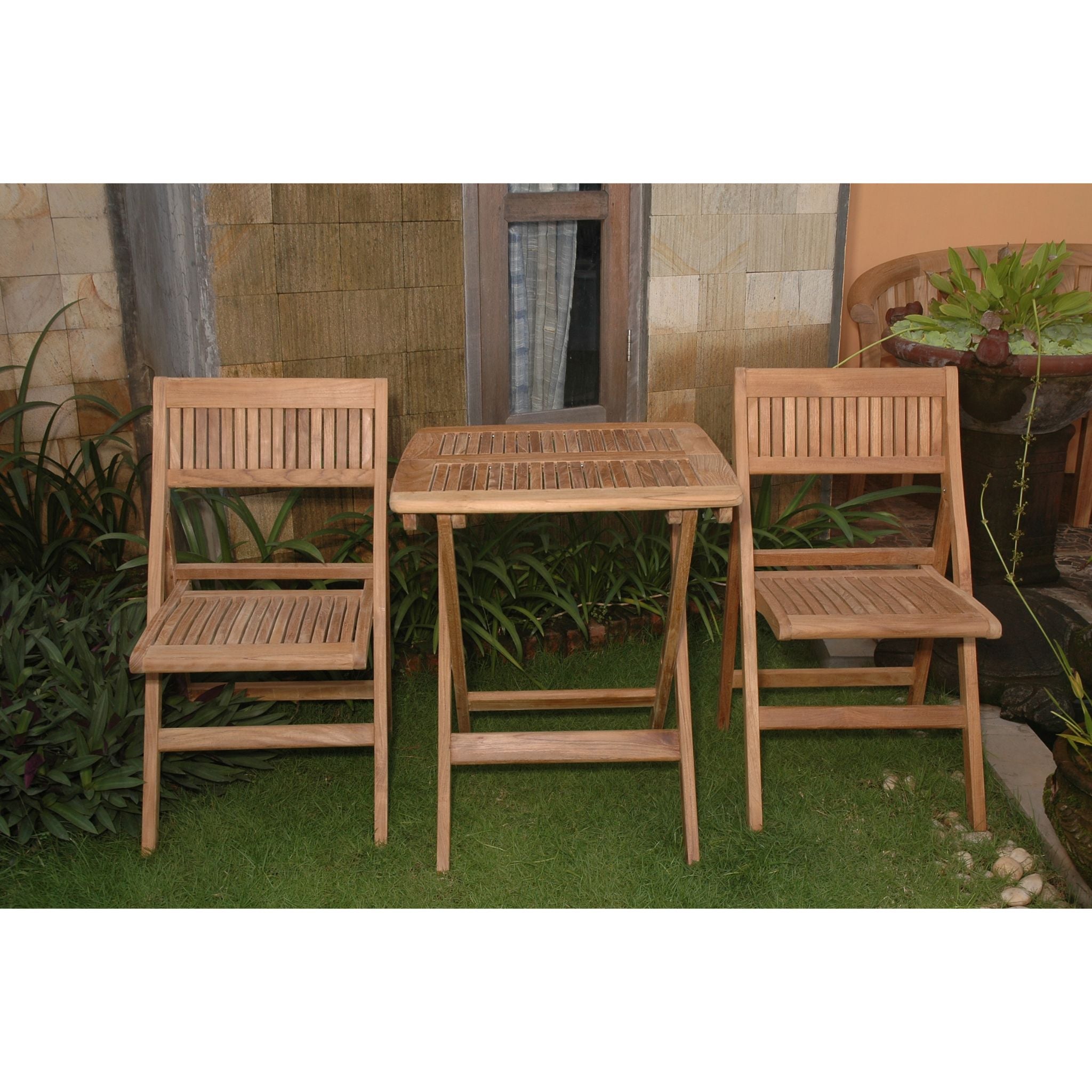 A three-piece outdoor wooden dining set consisting of two folding chairs and a small square folding table, placed on green grass in front of a stone wall and potted plants.