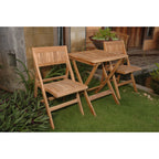 A three-piece outdoor patio set made of natural teak wood, including two folding chairs and a small square folding table, displayed on a green lawn in front of a building.