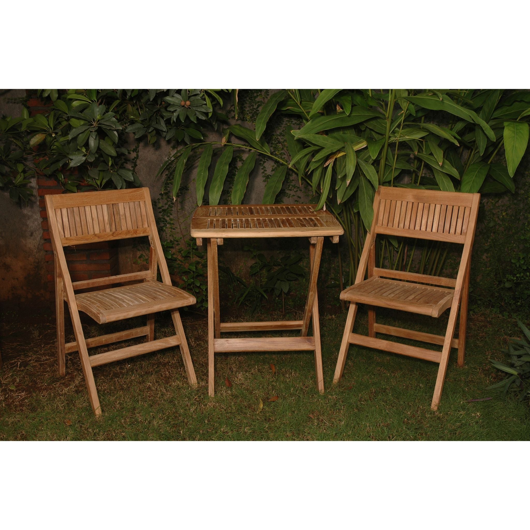 A three-piece outdoor wooden patio set consisting of two folding chairs and a small slatted side table, placed on grass in front of a leafy green wall.