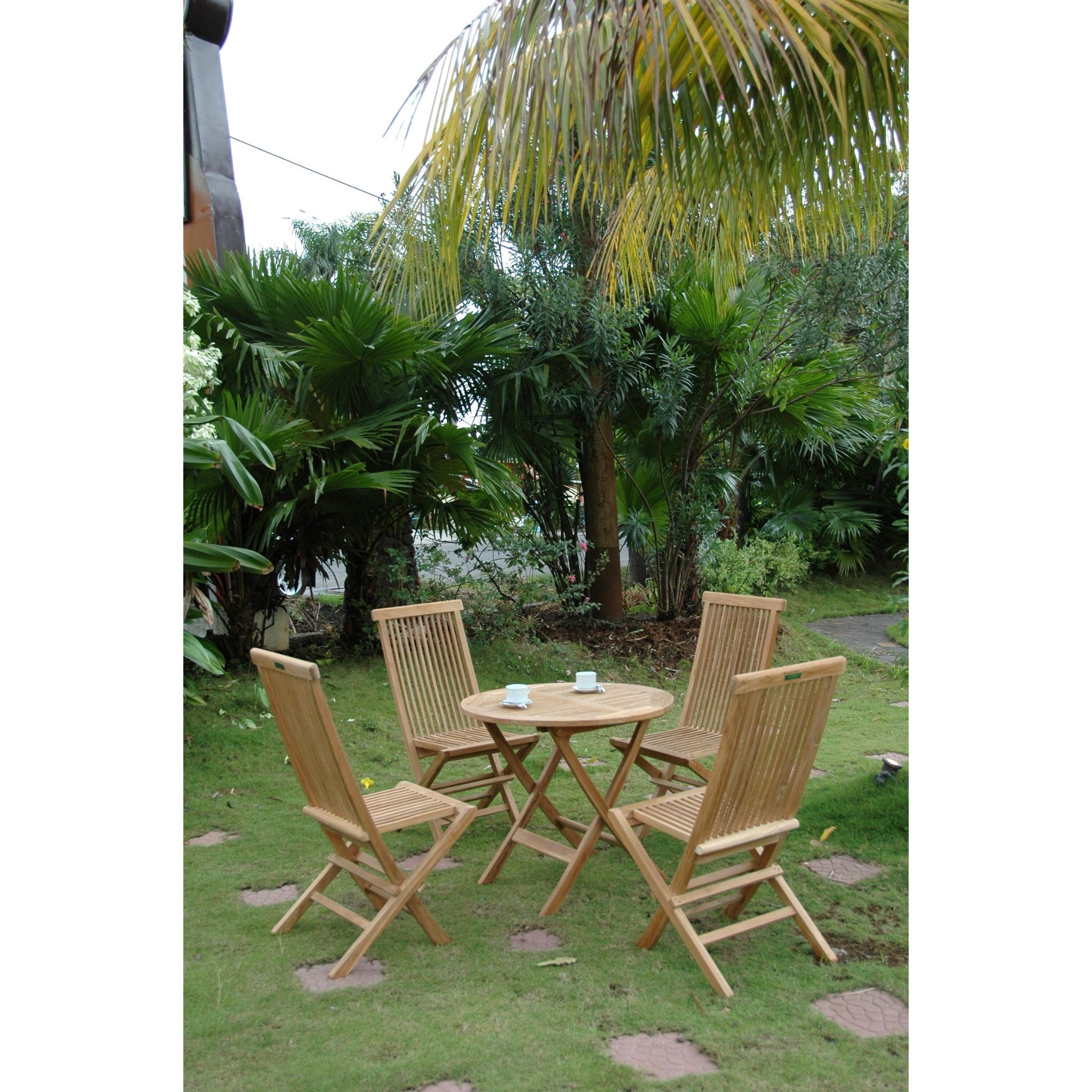 A four-piece outdoor wooden dining set consisting of a round folding table and four matching folding chairs, placed on a green lawn in a garden setting with palm trees and foliage in the background.