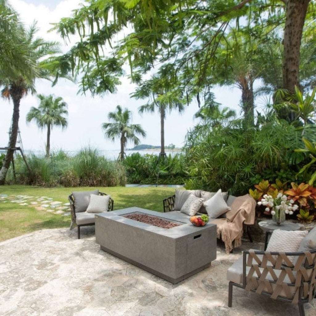 Outdoor patio furniture set in a tropical garden setting with palm trees and ocean view. The set includes a rectangular concrete fire pit table, a curved sectional sofa with gray cushions and a beige throw blanket, and two matching accent chairs on a stone patio.