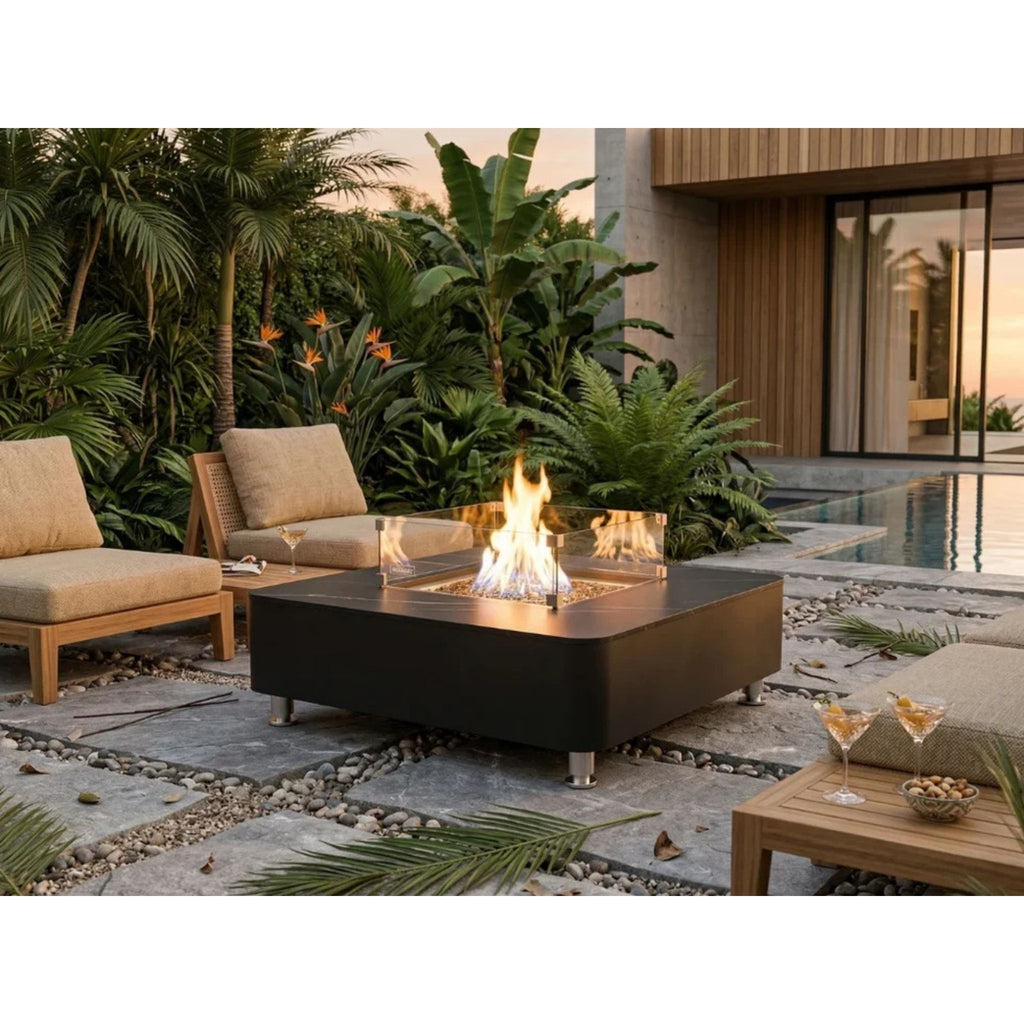 A modern outdoor patio scene with a black rectangular fire pit table in the center, surrounded by two beige lounge chairs, a small wooden side table with drinks, and tropical plants near a pool at dusk.