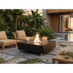 A modern outdoor patio scene with a black rectangular fire pit table in the center, surrounded by two beige lounge chairs, a small wooden side table with drinks, and tropical plants near a pool at dusk.