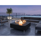 A modern outdoor patio scene at dusk with a black square fire pit table in the center, showing flames inside a clear glass wind guard. The patio is furnished with gray sectional sofas and armchairs, with a glass railing, pool, and city skyline in the background.