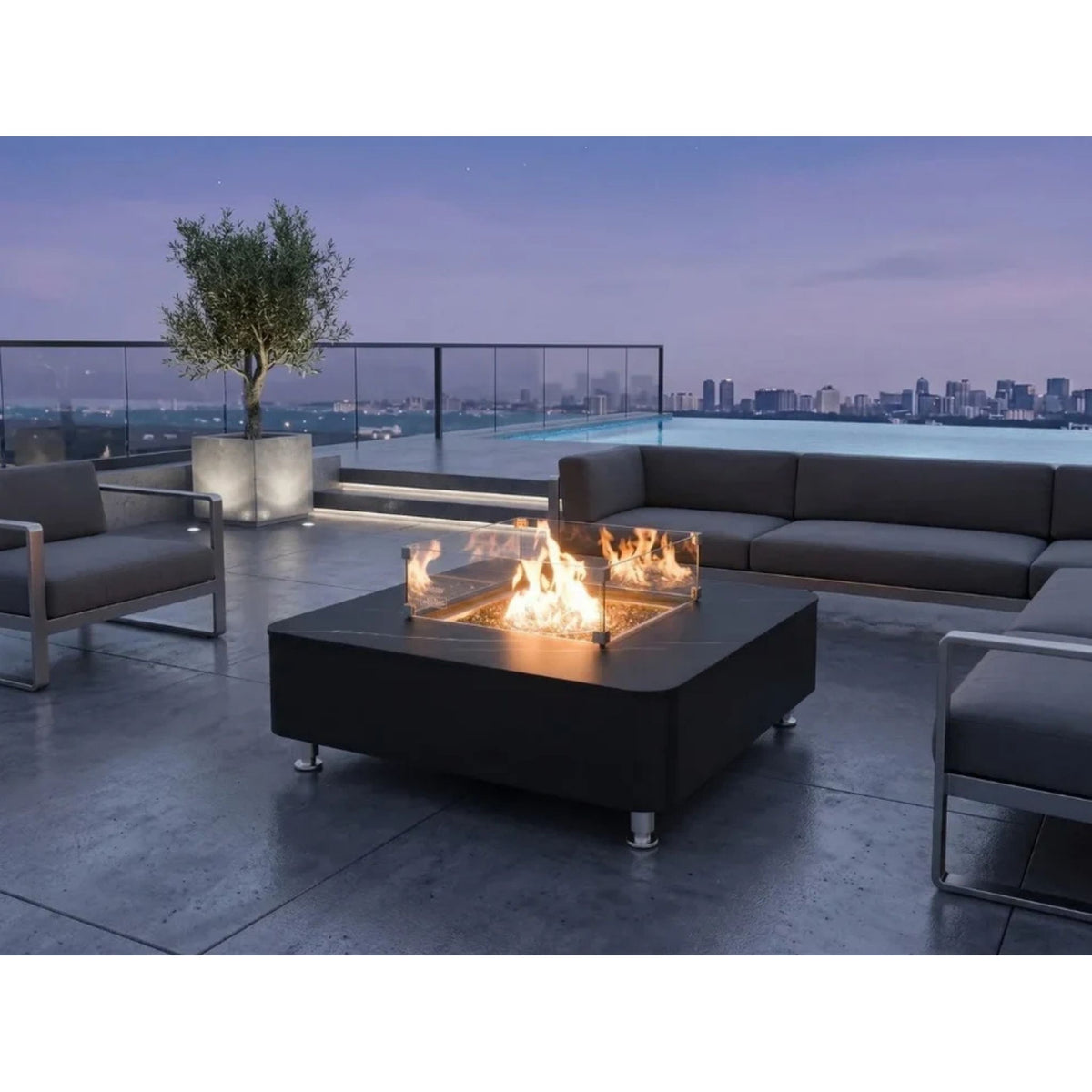 A modern outdoor patio scene at dusk with a black square fire pit table in the center, showing flames inside a clear glass wind guard. The patio is furnished with gray sectional sofas and armchairs, with a glass railing, pool, and city skyline in the background.