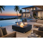 A modern outdoor fire pit table with a black metal frame and glass wind guard, lit with a warm fire, situated on a stone patio next to an infinity pool and a luxury coastal house at sunset.