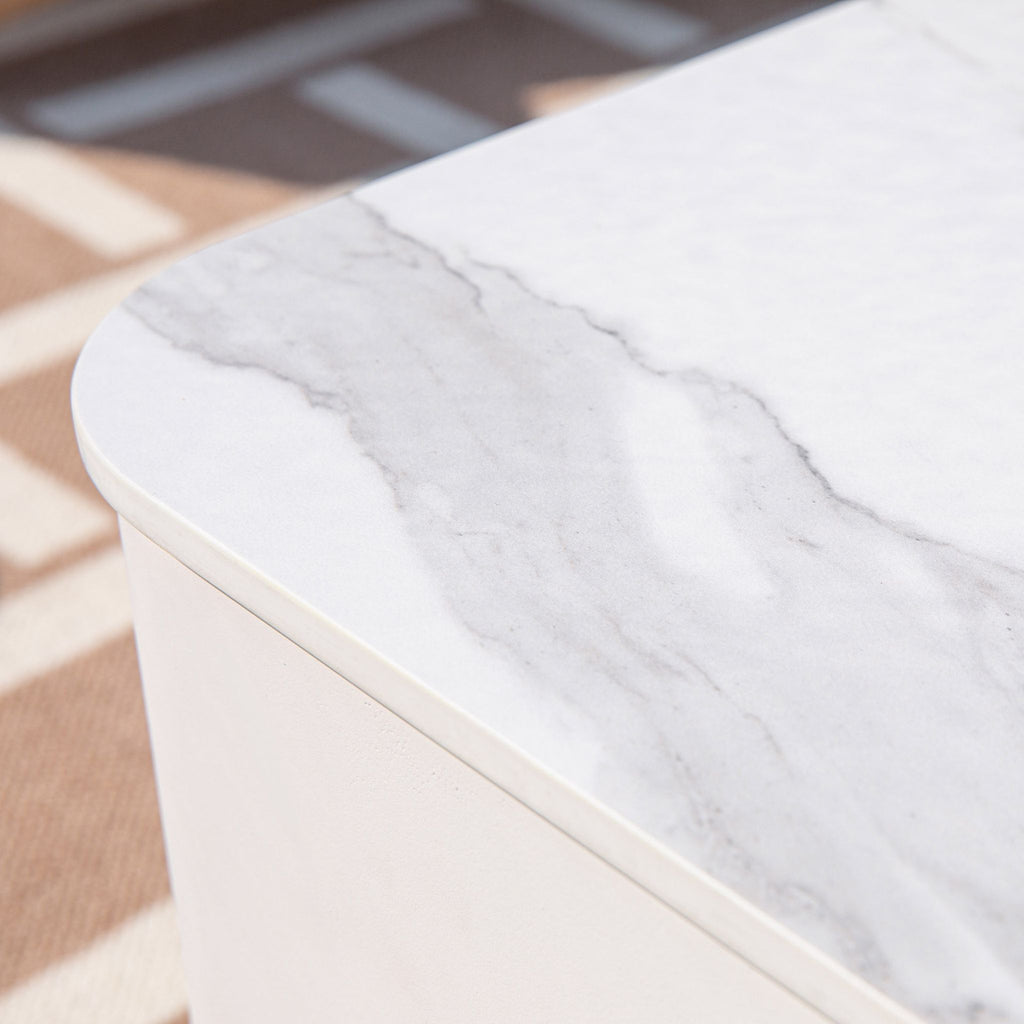 Close-up of a white marble-patterned tabletop corner with a smooth white base, photographed indoors on a patterned rug.