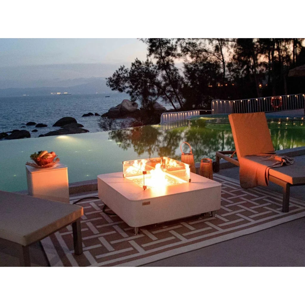 A modern outdoor fire pit table with a glowing fire, set on a patterned rug next to a swimming pool and ocean view at dusk.