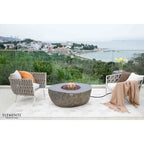 Outdoor patio furniture set featuring two woven gray armchairs and a round stone fire pit table on a light rug, overlooking a coastal landscape. The brand name ELEMENTI is visible in the bottom left corner.
