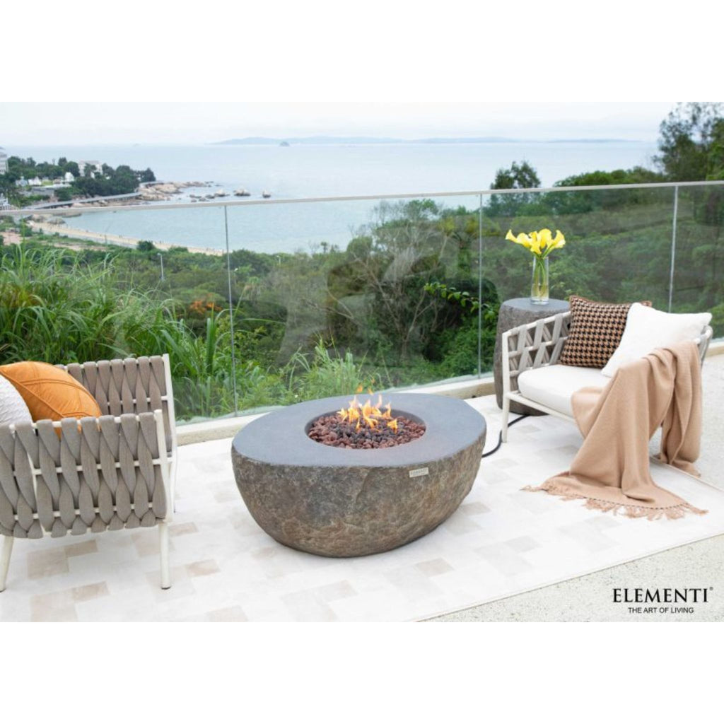 Outdoor patio scene with a round stone fire pit table in the center, flanked by two woven armchairs and a throw blanket, overlooking a coastal landscape. The brand name Elementi appears in the bottom right corner.