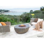 Outdoor patio scene with a round stone fire pit table in the center, flanked by two woven armchairs and a throw blanket, overlooking a coastal landscape. The brand name Elementi appears in the bottom right corner.