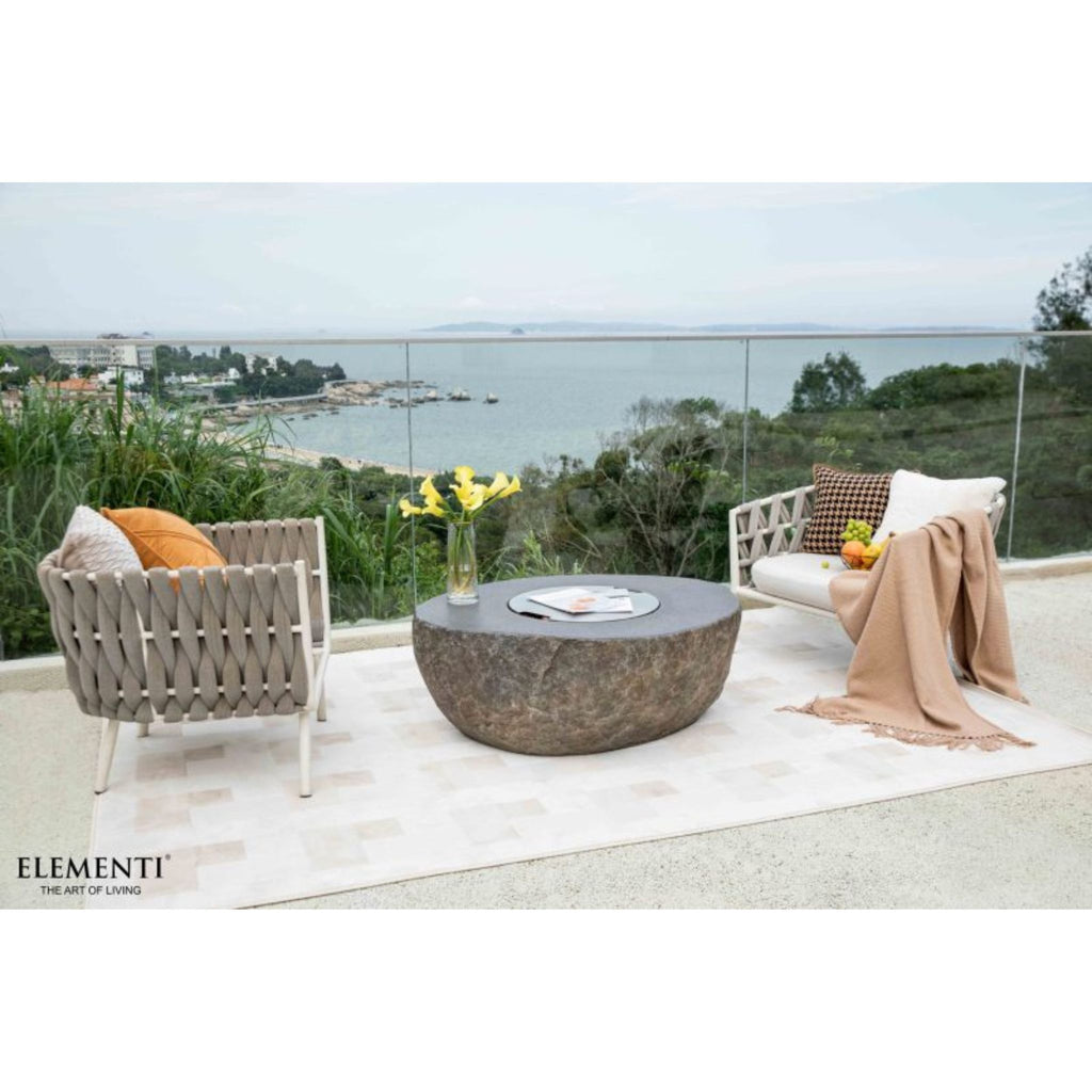 Outdoor patio furniture set featuring a woven gray armchair, a matching armchair with a draped beige throw, and a round stone coffee table on a light rug, overlooking a scenic coastal view. The brand name ELEMENTI is visible in the bottom left corner.