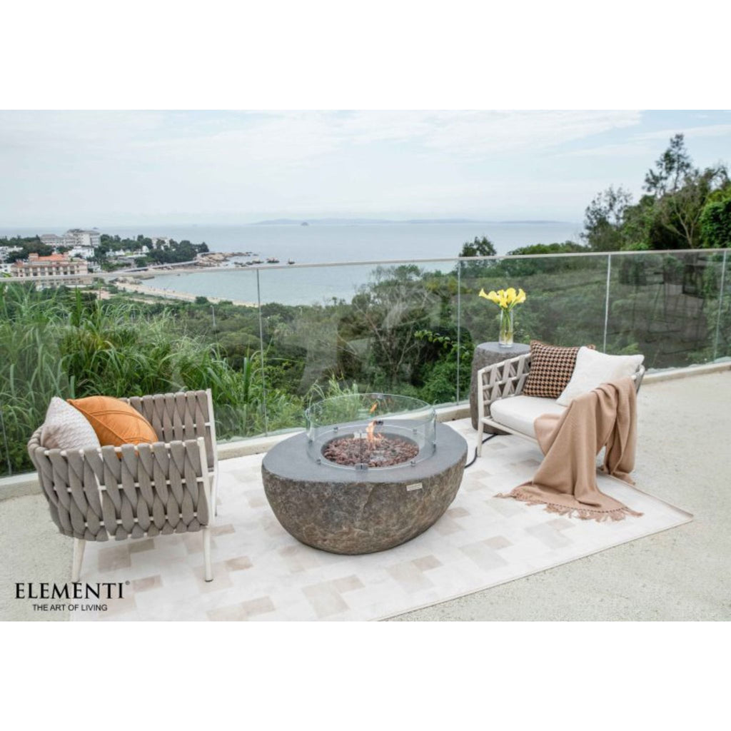 Outdoor patio furniture set on a terrace overlooking a coastal landscape, featuring a round stone fire pit table, two woven armchairs with cushions, a side table with yellow flowers, and a draped throw blanket. The brand name Elementi is visible in the bottom left corner.