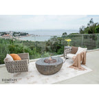 Outdoor patio furniture set on a terrace overlooking a coastal landscape, featuring a round stone fire pit table, two woven armchairs with cushions, a side table with yellow flowers, and a draped throw blanket. The brand name Elementi is visible in the bottom left corner.