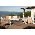 Outdoor patio furniture set on a wooden deck overlooking the ocean, featuring a cream woven sofa with patterned pillows, two matching armchairs, a round stone fire pit, and a thatched cabana with a draped throw blanket. The brand name ELEMENTI is visible in the bottom right corner.