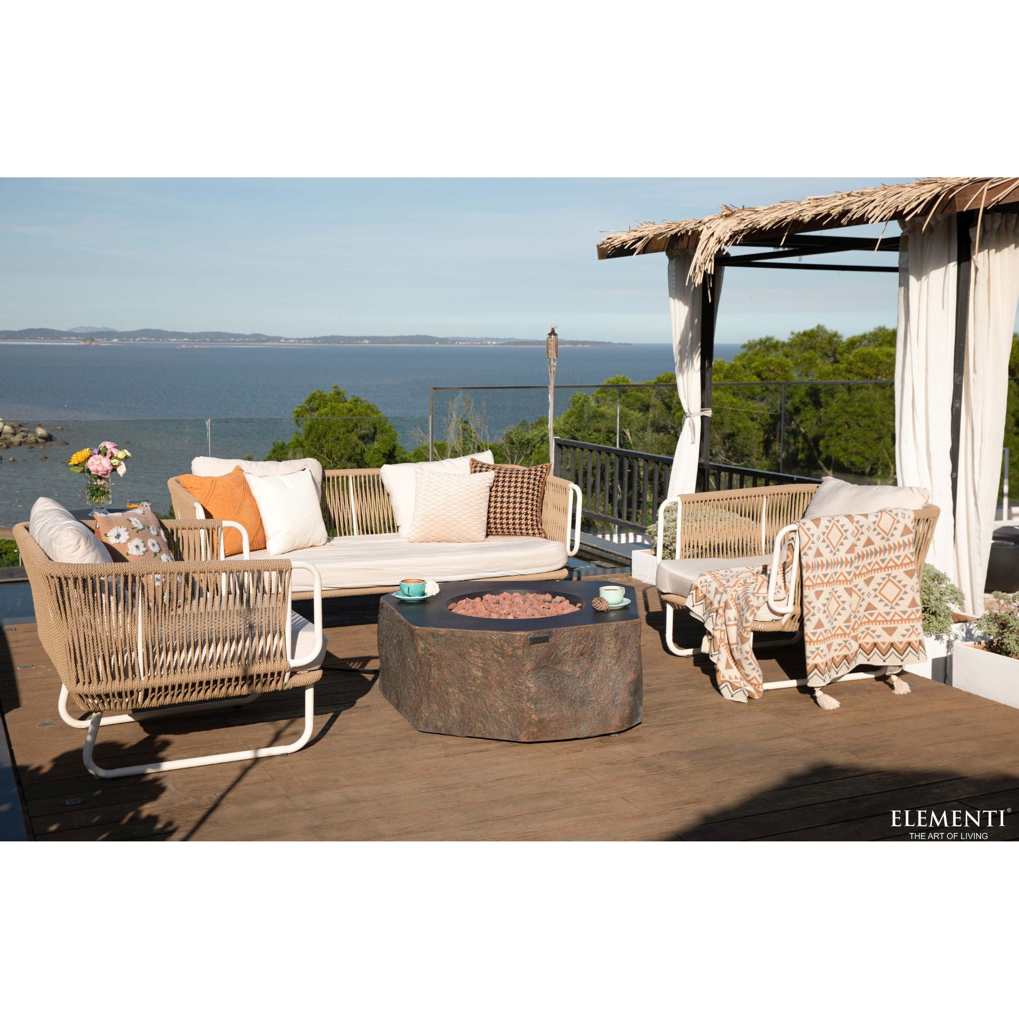 Outdoor patio furniture set on a wooden deck overlooking the ocean, featuring a cream woven sofa with patterned pillows, two matching armchairs, a round stone fire pit, and a thatched cabana with a draped throw blanket. The brand name ELEMENTI is visible in the bottom right corner.