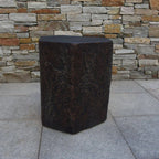 A dark brown hexagonal stone stool with a textured, weathered surface, set on a light gray tiled floor in front of a rustic stone wall.