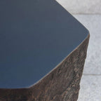 Close-up of a dark charcoal gray metal table top with a rustic, rough-hewn dark brown stone base, set against a light gray tiled floor.