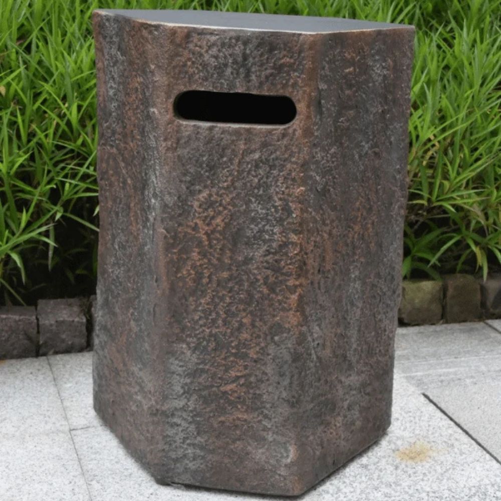 A rustic, hexagonal stone-look trash can or planter with a textured dark brown and gray finish, featuring a horizontal slot cutout near the top, set outdoors on a paved surface with green foliage in the background.