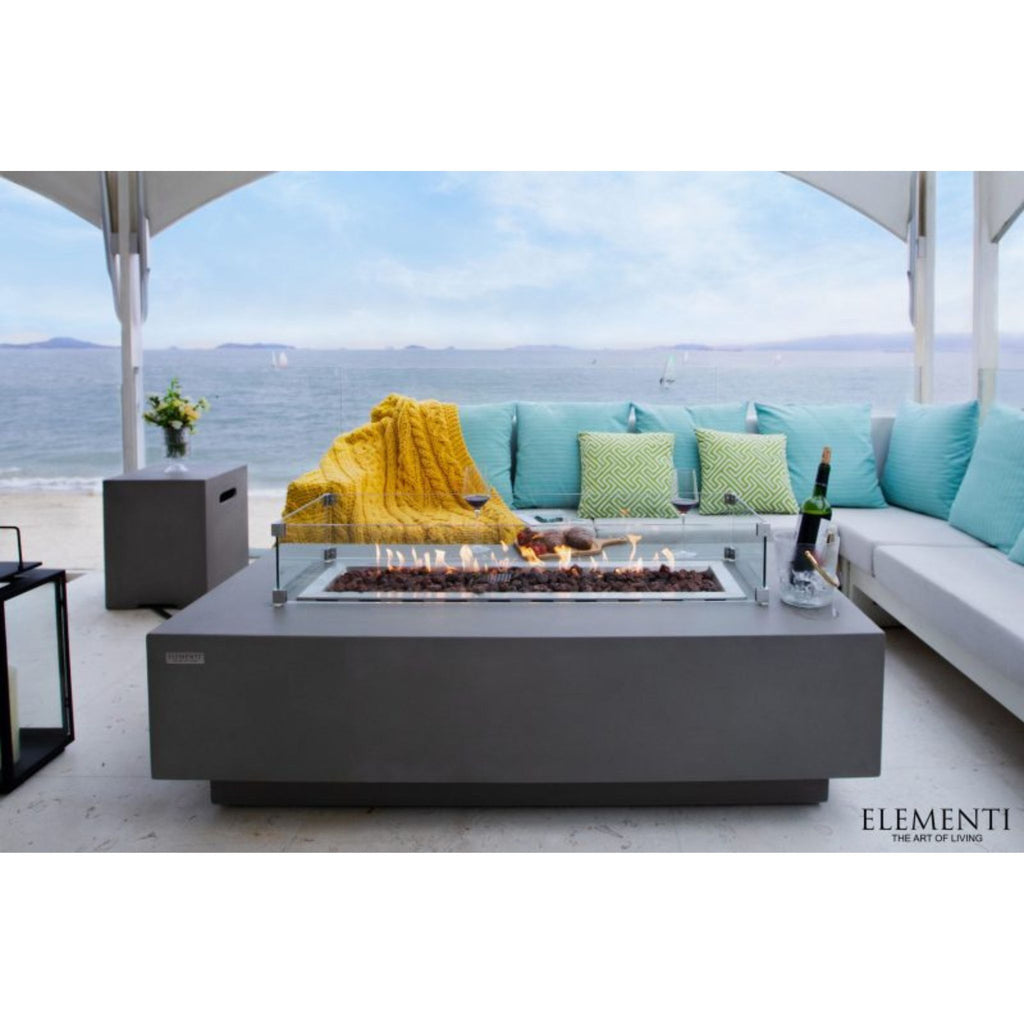 Elementi outdoor rectangular fire pit table with a glass wind guard, set on a patio next to a modern sectional sofa with turquoise pillows and a yellow throw blanket, overlooking the ocean.