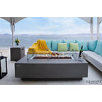 Elementi outdoor rectangular fire pit table with a glass wind guard, set on a patio next to a modern sectional sofa with turquoise pillows and a yellow throw blanket, overlooking the ocean.