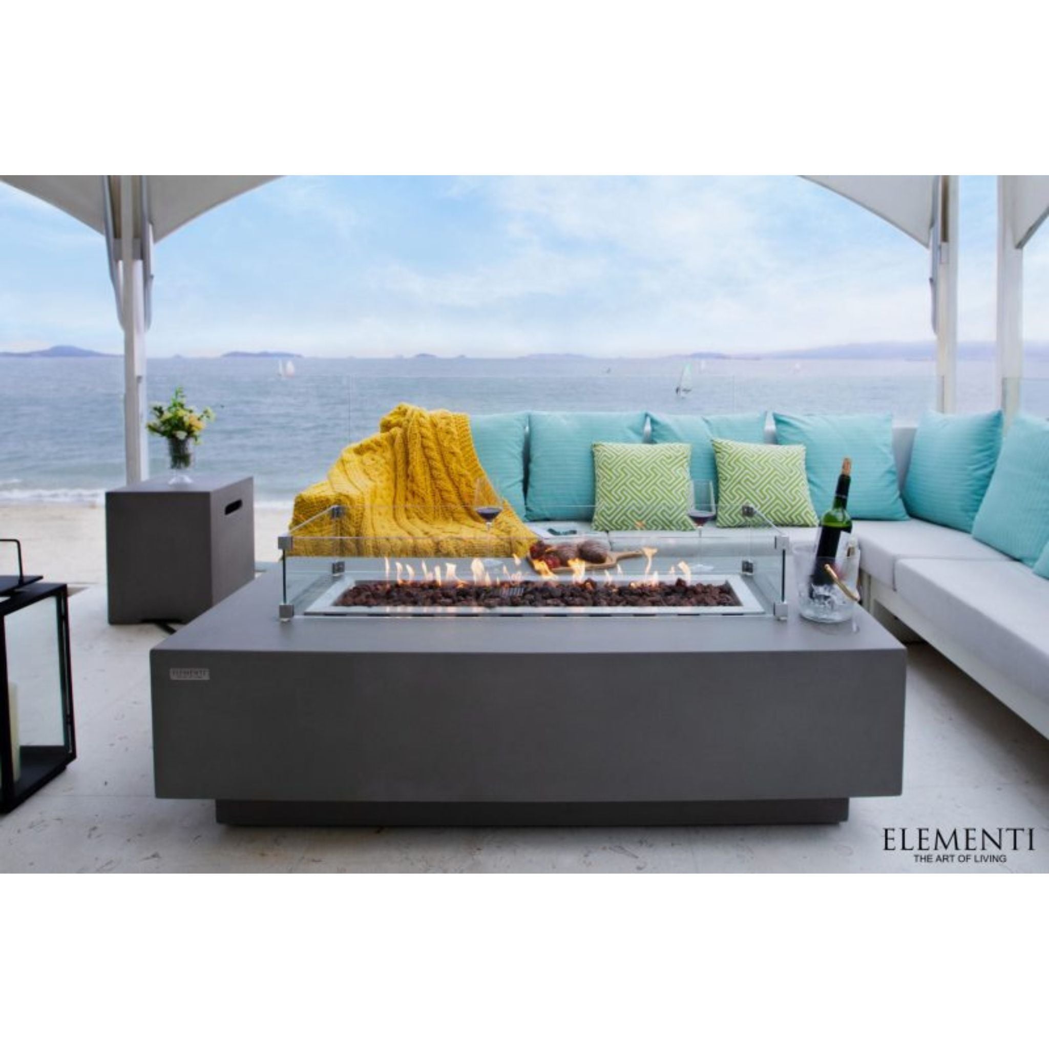 Elementi outdoor rectangular fire pit table with a glass wind guard, set on a patio next to a modern sectional sofa with turquoise pillows and a yellow throw blanket, overlooking the ocean.