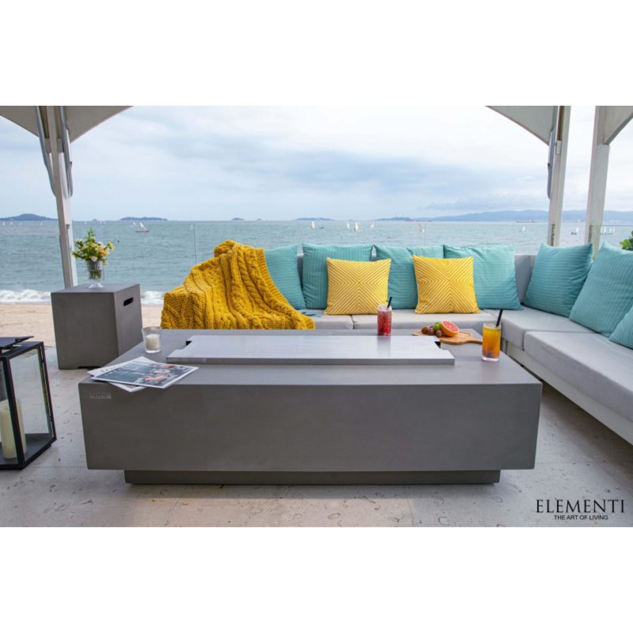 A modern outdoor patio scene by the beach with a large gray rectangular coffee table in the foreground, a sectional sofa with teal and yellow pillows, and an ocean view under a canopy. The brand name ELEMENTI appears in the bottom right corner.