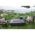 Outdoor patio furniture set on a grassy terrace overlooking a coastal landscape, featuring a rectangular fire pit table, two dark armchairs with patterned cushions, and a small side table with pink flowers. The brand name Elementi is visible in the bottom left corner.