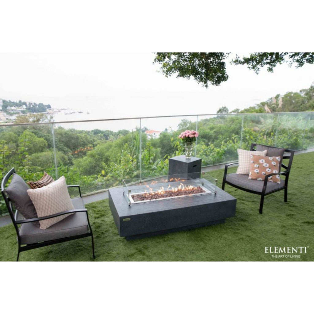 Outdoor patio furniture set on a green lawn with a glass railing and forest view, featuring two dark gray armchairs with patterned pillows and a rectangular black fire pit table. The brand name Elementi is visible in the bottom right corner.