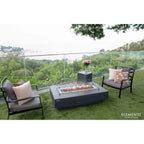 Outdoor patio furniture set on a green lawn with a glass railing and forest view, featuring two dark gray armchairs with patterned pillows and a rectangular black fire pit table. The brand name Elementi is visible in the bottom right corner.