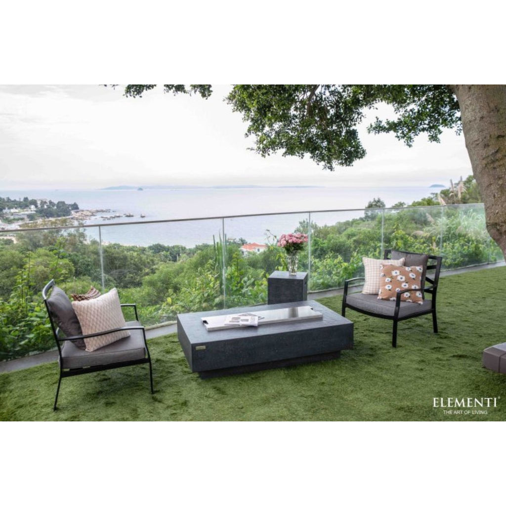 Outdoor patio furniture set on green artificial turf overlooking a scenic ocean view. The set includes two black-framed armchairs with gray cushions and patterned pillows, a rectangular dark gray coffee table, and a small matching side table with a vase of pink flowers.