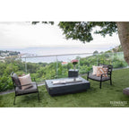 Outdoor patio furniture set on green artificial turf overlooking a scenic ocean view. The set includes two black-framed armchairs with gray cushions and patterned pillows, a rectangular dark gray coffee table, and a small matching side table with a vase of pink flowers.