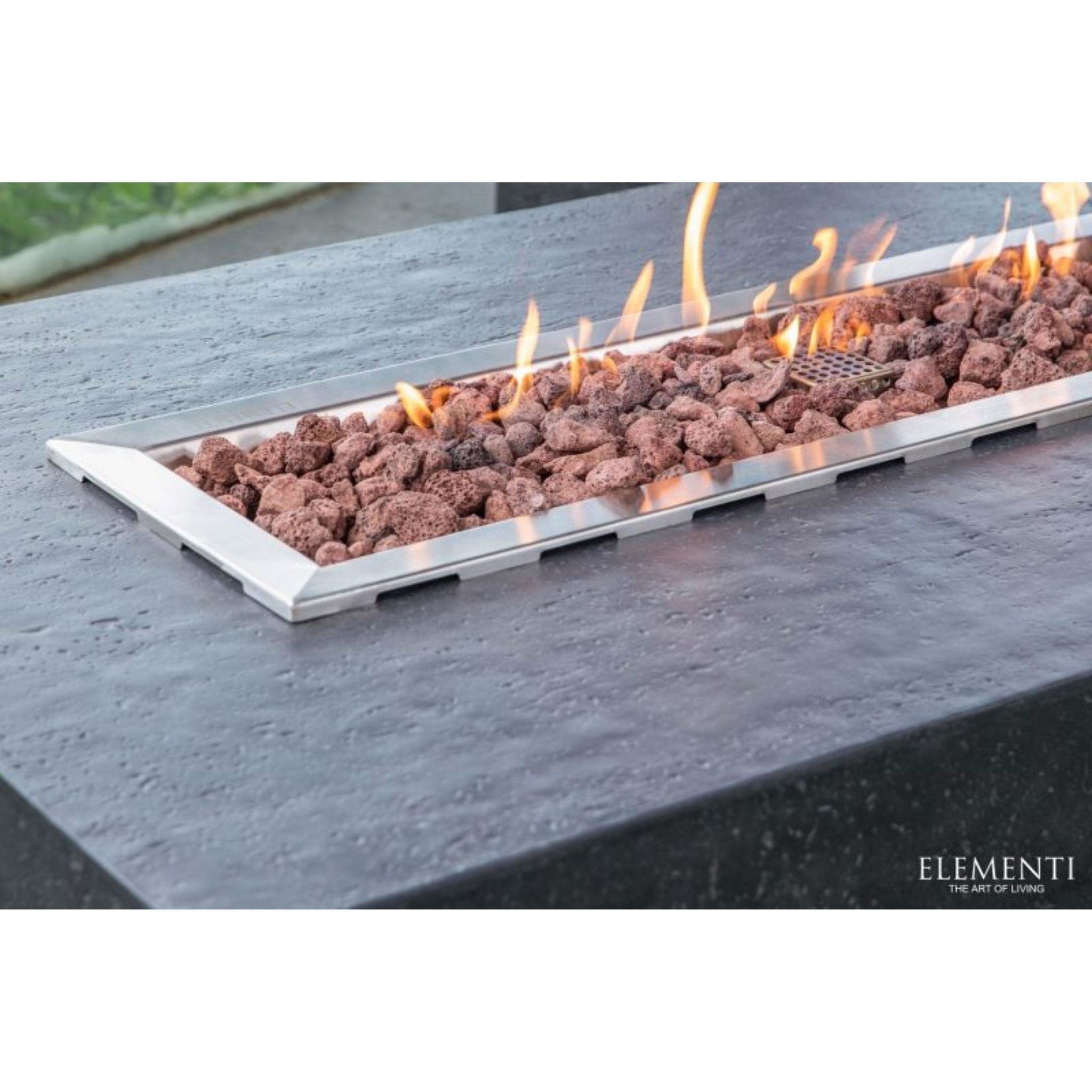 Elementi outdoor rectangular fire pit with a stainless steel frame, filled with brown lava rocks and active orange flames, set into a dark stone table.