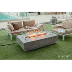 Outdoor patio furniture set including a rectangular concrete fire pit table with glass wind guard, a beige cushioned sofa, and a matching armchair on green artificial turf, with the Elementi logo in the bottom right corner.