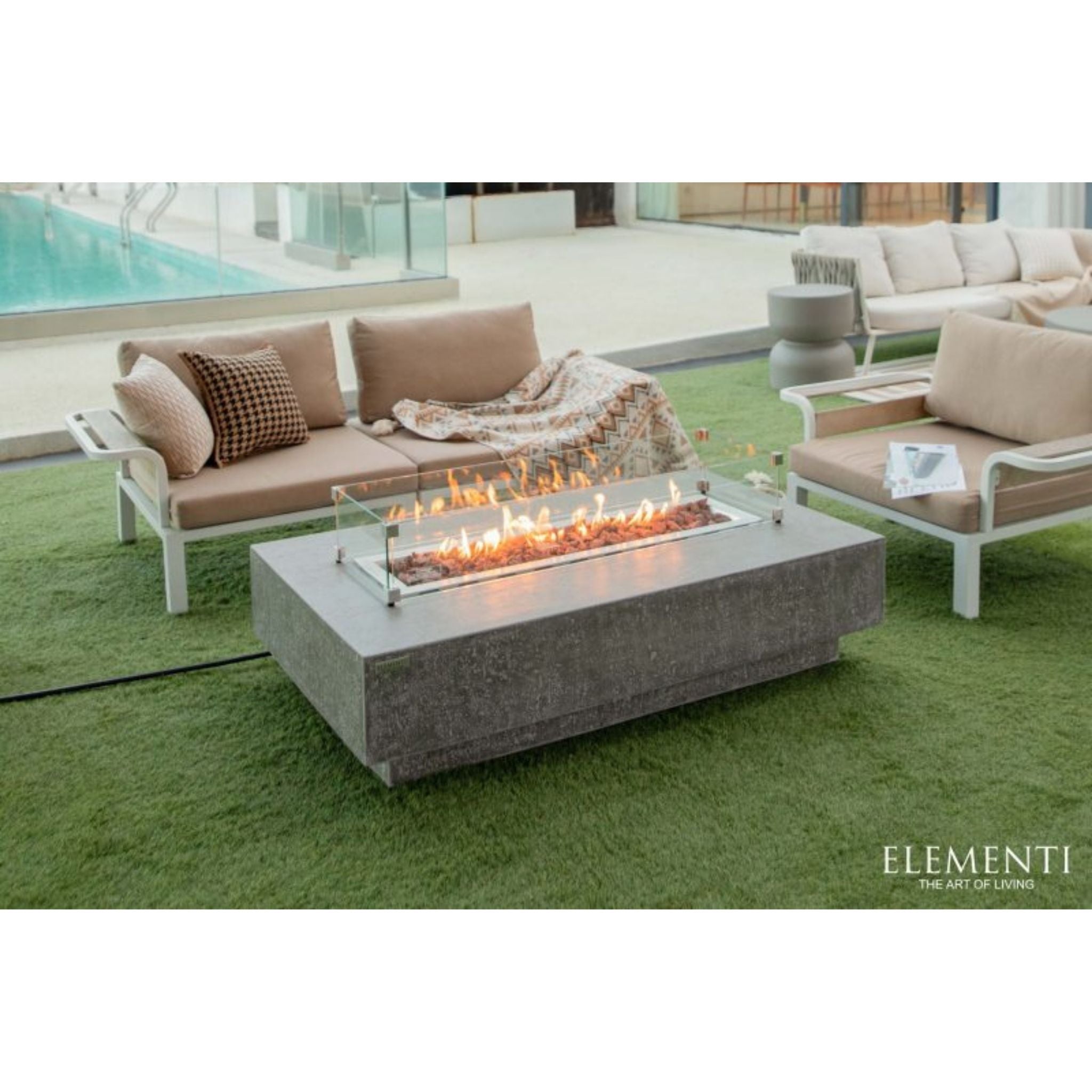 Outdoor patio furniture set including a rectangular concrete fire pit table with glass wind guard, a beige cushioned sofa, and a matching armchair on green artificial turf, with the Elementi logo in the bottom right corner.