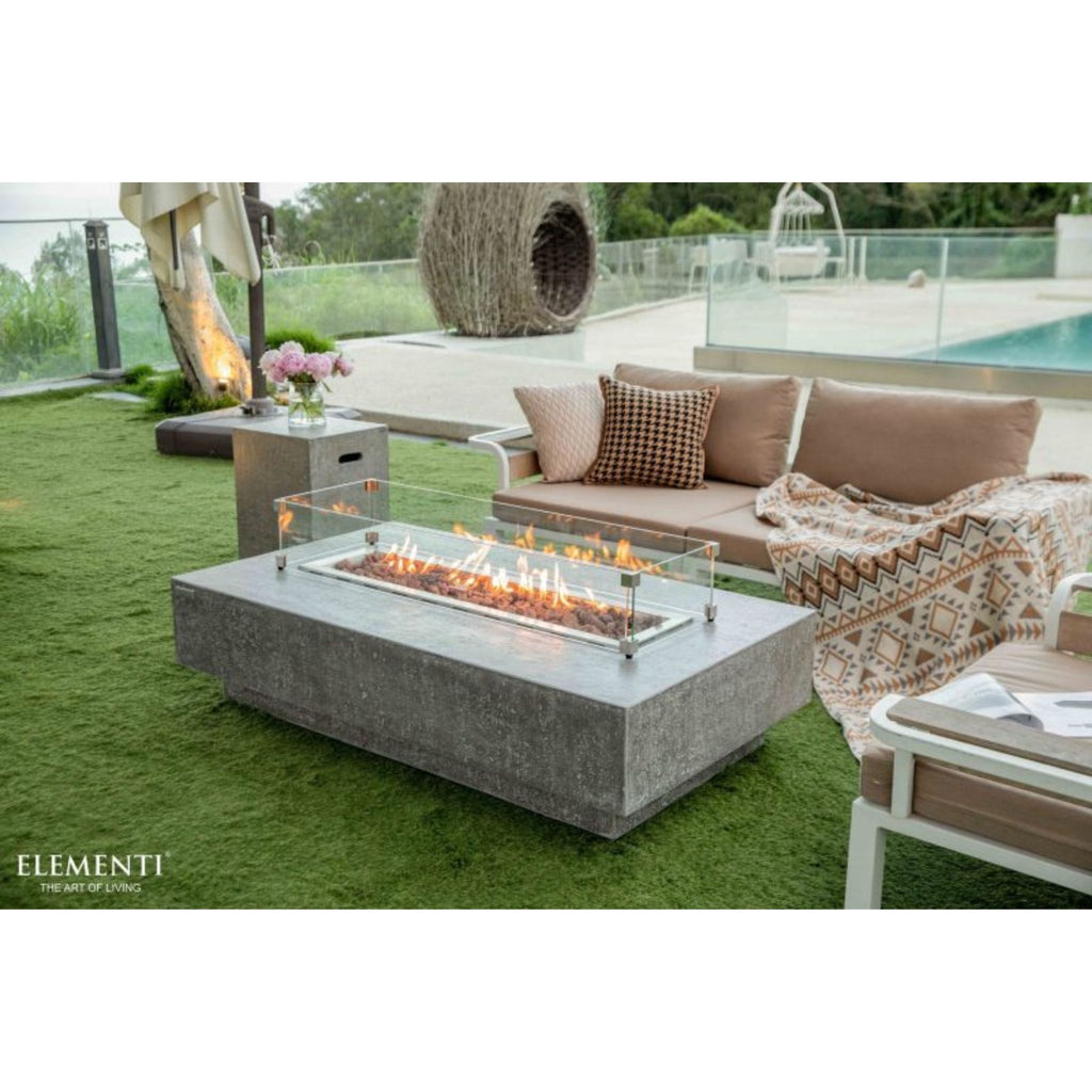 Outdoor patio scene with a rectangular concrete fire pit table in the foreground, a beige sectional sofa with pillows and a patterned throw, and a pool area in the background. The brand name ELEMENTI appears in the lower left corner.