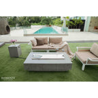 Outdoor patio furniture set on green artificial grass beside a swimming pool, featuring a rectangular concrete-look coffee table, a beige cushioned sofa with pillows and a throw blanket, a matching armchair, and a small side table. The brand name ELEMENTI is visible in the bottom left corner.