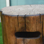 Close-up of a rustic wooden stump stool with a natural tree-ring top and a cutout handle on the front, photographed outdoors with grass and a building in the background.