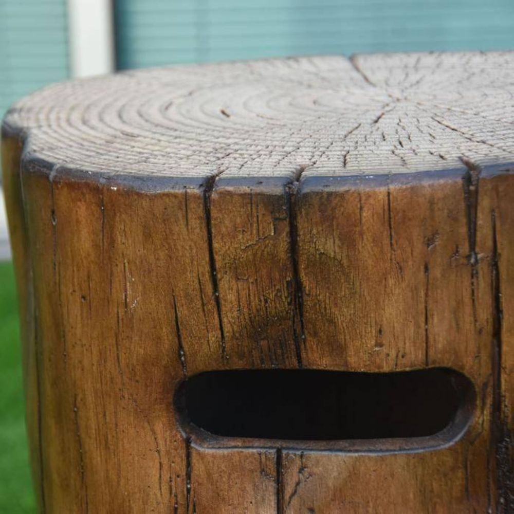 Close-up of a rustic wooden stump stool with a natural tree-ring top and a cutout handle on the front, photographed outdoors with grass and a building in the background.