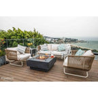 Outdoor patio furniture set on a wooden deck overlooking the ocean, featuring a wicker sofa, two matching armchairs with striped and geometric pillows, and a dark stone fire pit table.
