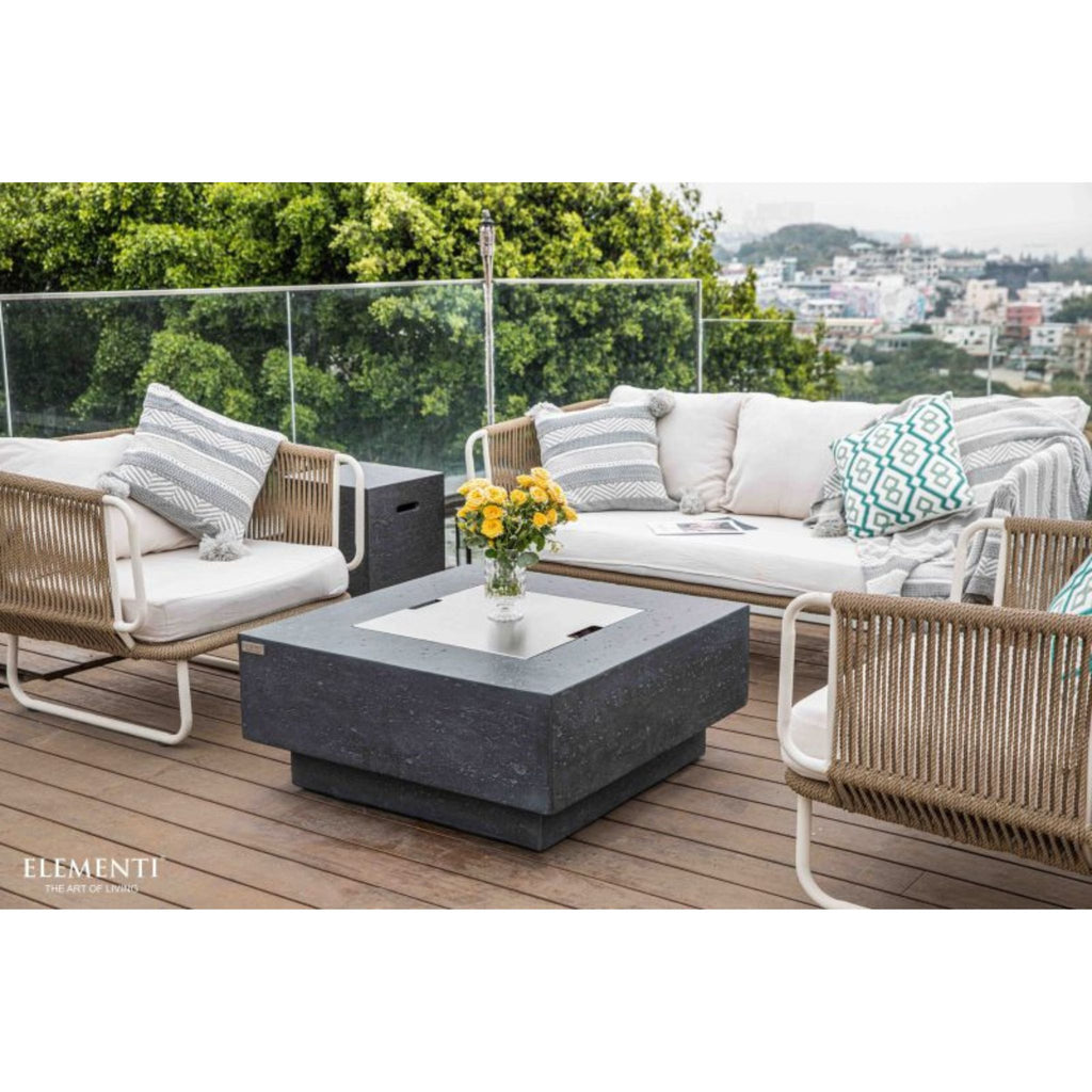 Outdoor patio furniture set on a wooden deck with a glass railing, featuring a dark gray square fire pit table, a white and beige woven sofa, two matching armchairs, and decorative pillows. Lush green trees and a distant cityscape are visible in the background.
