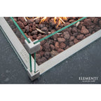 Close-up of an Elementi outdoor fire pit with a silver metal frame, clear glass panels, and lava rocks with visible flames, on a dark textured surface.