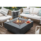 A modern outdoor patio scene featuring a square Elementi fire pit table with a dark gray stone-look body, clear glass wind guard, and visible flames. The table is styled on a wooden deck between two white outdoor sofas with gray and white patterned cushions.