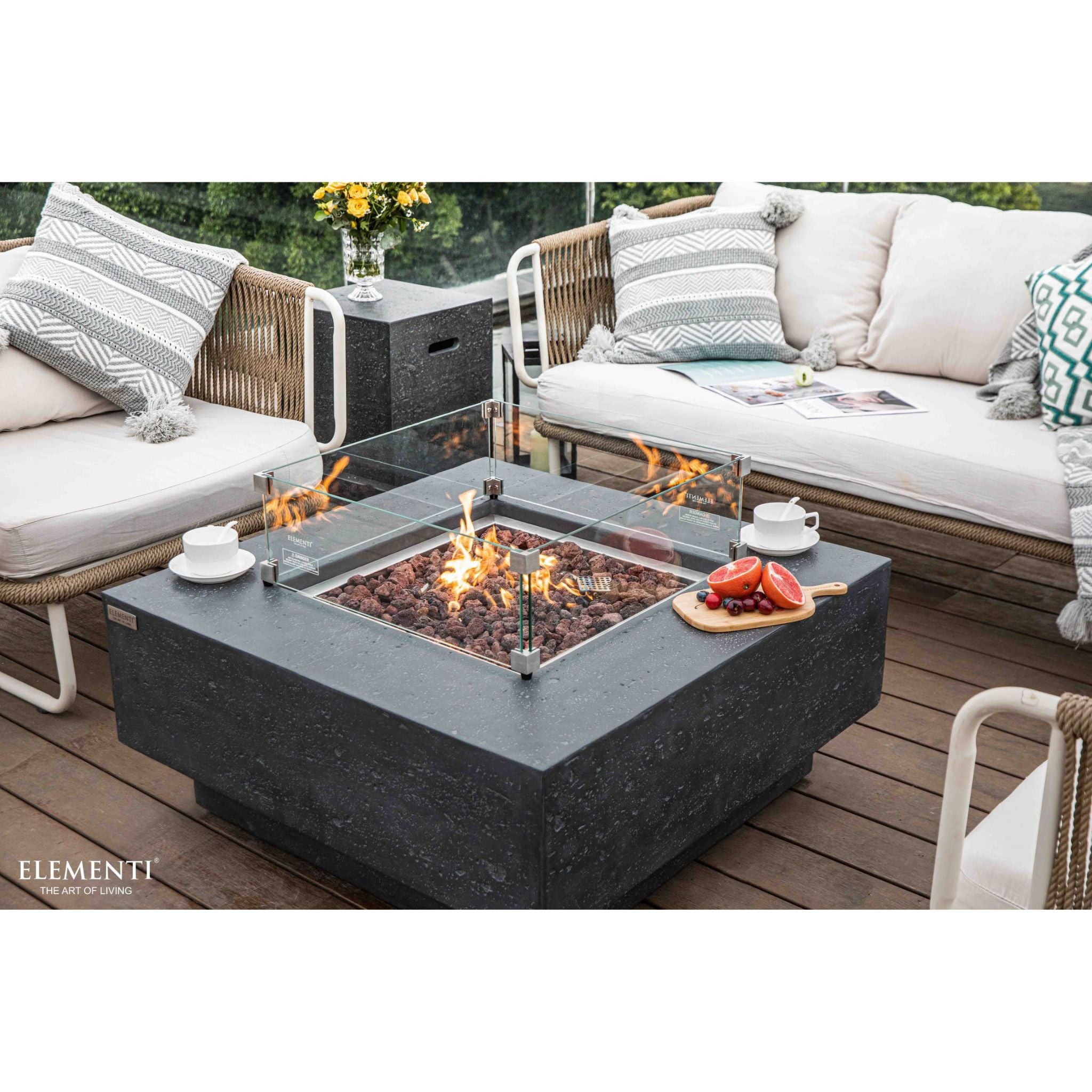 A modern outdoor patio scene featuring a square Elementi fire pit table with a dark gray stone-look body, clear glass wind guard, and visible flames. The table is styled on a wooden deck between two white outdoor sofas with gray and white patterned cushions.