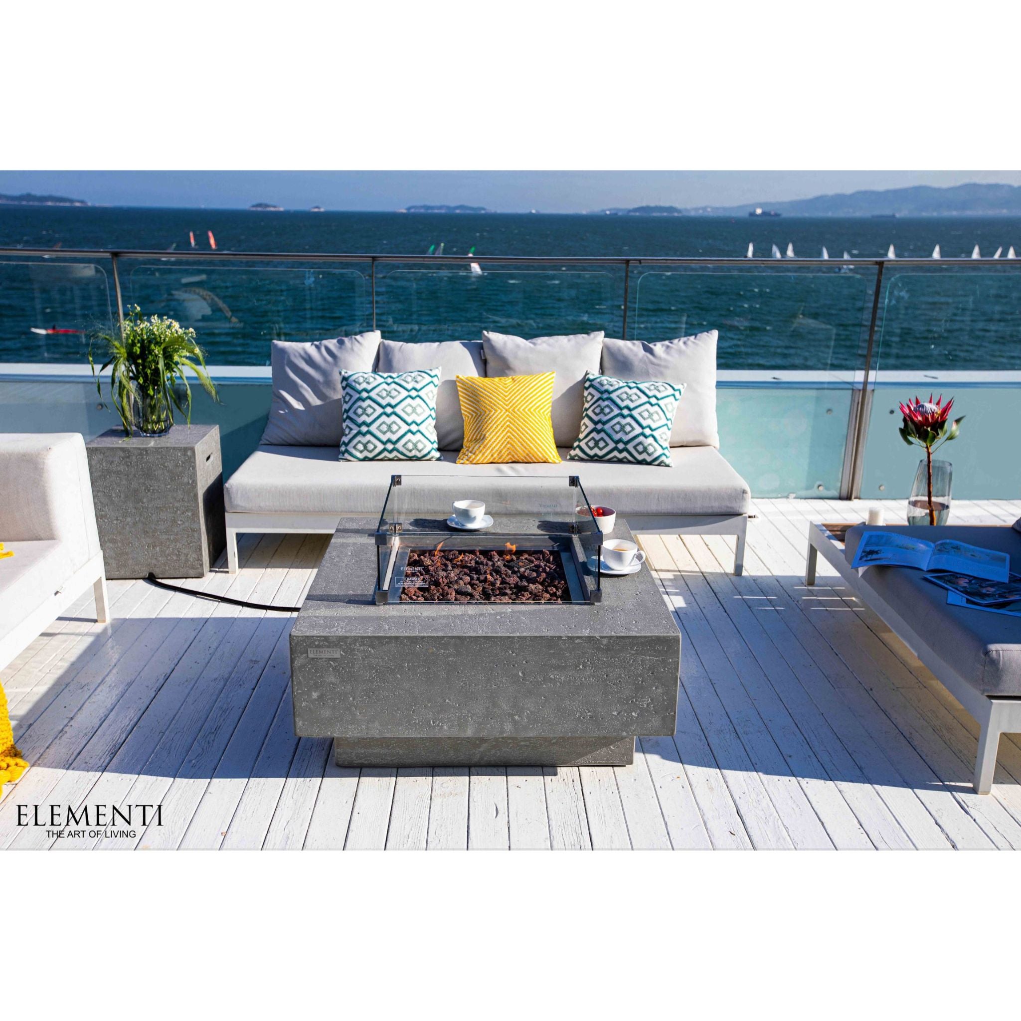 Outdoor patio furniture set on a white-decked terrace overlooking the ocean, featuring a gray concrete fire pit table, a light gray sectional sofa with blue and yellow accent pillows, and a matching lounge chair. The brand name ELEMENTI is visible in the bottom left corner.